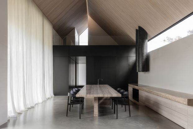 A refined open-plan dining and living space with a long timber dining table and black chairs beneath a dramatic pitched, timber-lined ceiling. Full-height sheer curtains line the glazed wall, while dark built-in cabinetry and a substantial timber-lined media or storage console create a strong contrast against the pale concrete floor and light walls. The room has a spacious, minimalist character with a prominent black structural beam and wall-mounted fireplace or dark feature element.