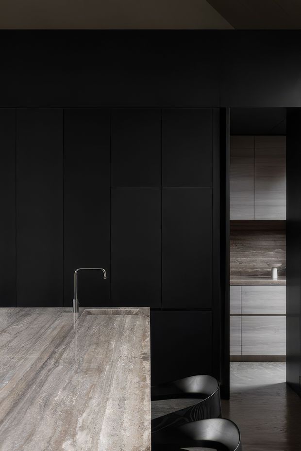 A moody, minimalist kitchen interior dominated by floor-to-ceiling matte black cabinetry. A large pale grey stone island with an integrated sink and slim chrome tap occupies the foreground, accompanied by sculptural black bar stools. Through the opening at right, additional pale timber cabinetry, a stone work surface, and dark timber flooring are visible. The space has a refined, high-contrast monochromatic palette and subdued lighting.