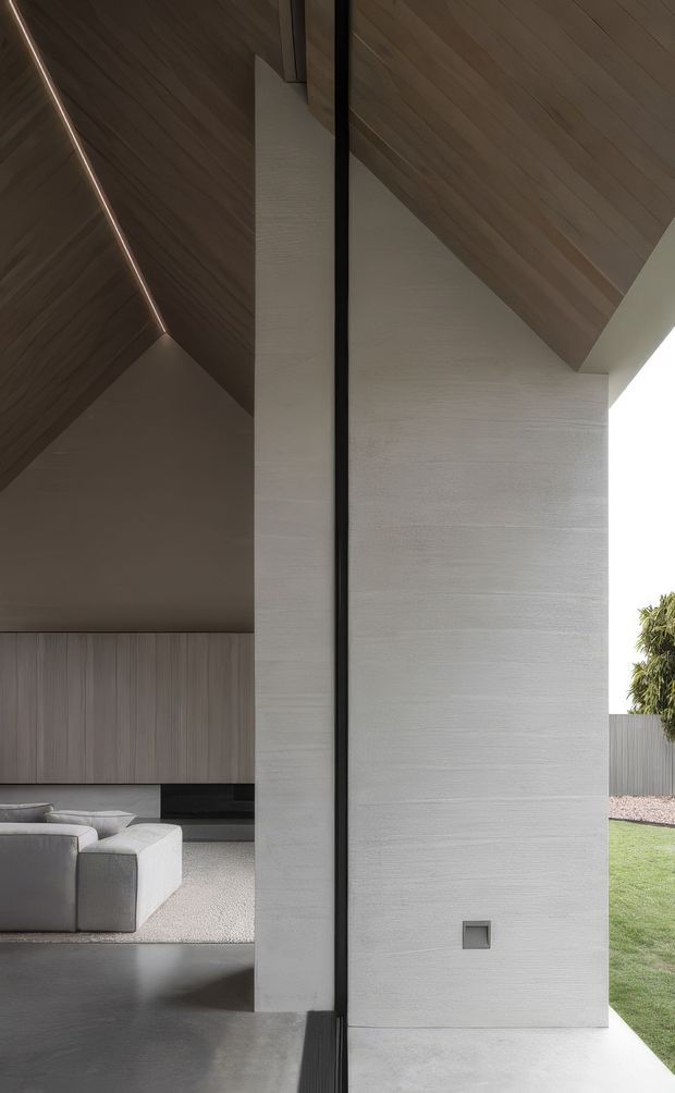 A refined modern architectural interior viewed through a large open glazed corner or doorway. The composition features a tall textured light-grey wall, dark slender framing, a sharply angled timber-lined ceiling with concealed linear lighting, polished dark flooring, and a partially visible minimalist living room with pale modular seating. A manicured lawn and planting are visible outside.