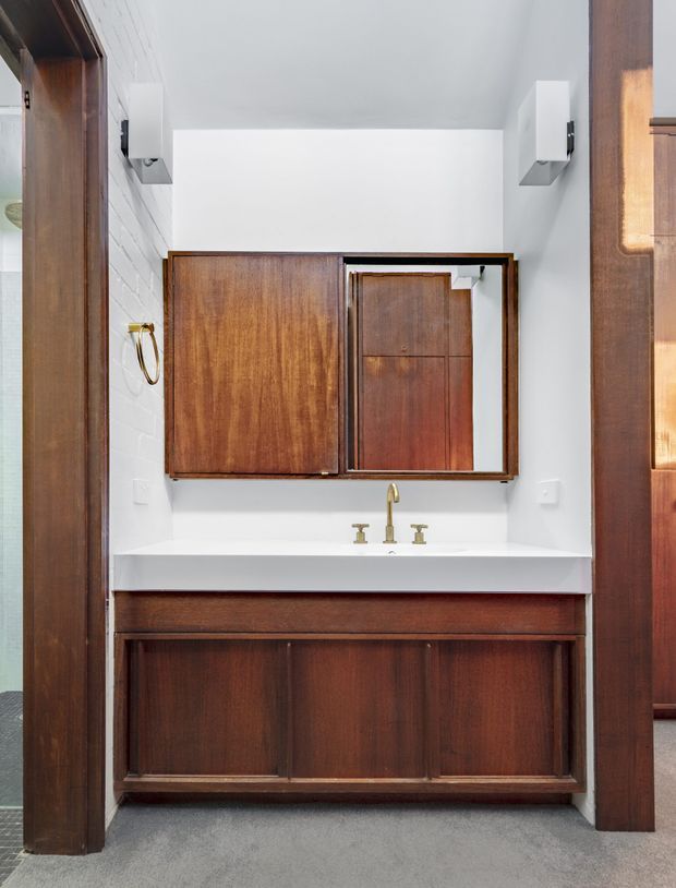 A compact bathroom vanity area with dark timber cabinetry, a white integrated benchtop and basin, brass tapware, wall-mounted mirrored storage, white painted brick walls, and grey flooring. The image is framed from the bathroom, with timber door or trim elements visible at both sides.