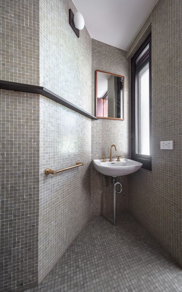 A compact contemporary bathroom with grey mosaic-tiled walls and floor, a wall-mounted white ceramic basin, exposed brass-toned plumbing and tapware, a narrow black-framed window, a timber-framed mirror, and a dark floating shelf. The space is tall and narrow with a clean, refined material palette.