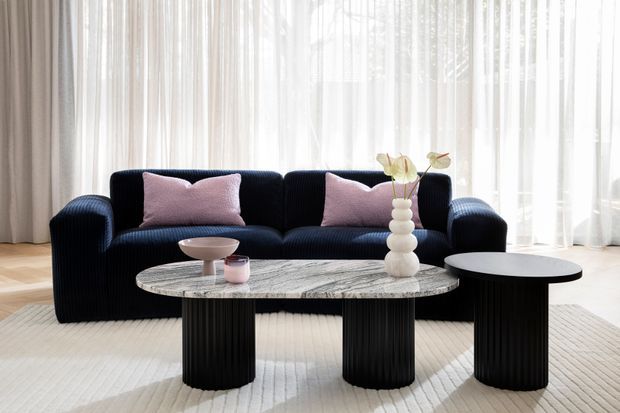 A bright, refined living room with a dark ribbed sectional sofa, blush pink cushions, and sheer floor-to-ceiling curtains. A large oval marble coffee table with sculptural black fluted legs is paired with a smaller round black side table. Pale timber flooring and a light textured rug create a soft, neutral setting, with a minimalist floral arrangement as an accent.