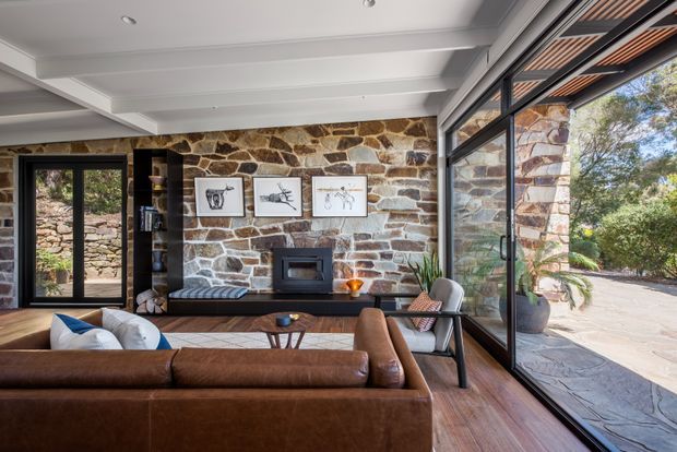 A warm, rustic-modern living room with a large brown leather sectional facing a stone feature wall and compact fireplace. The room has extensive black-framed glazing and doors, exposed natural stone masonry, timber flooring, and a white coffered or exposed-beam ceiling. A landscaped outdoor area is visible through the glazing.