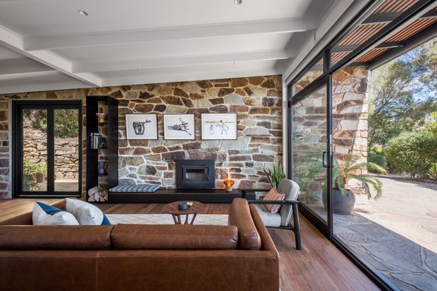 A warm, contemporary living room with a large brown leather sectional sofa, timber flooring, and a prominent natural rough-stone feature wall with a built-in fireplace. Black-framed full-height glazing opens toward a landscaped outdoor area, while exposed white ceiling beams and a timber-lined ceiling recess add architectural character. Minimal artwork, shelving, and potted greenery complete the refined interior.