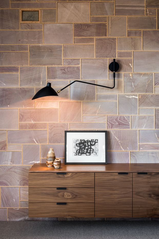 Close-up interior view of a refined mid-century-inspired living or dining space, featuring a dramatic wall clad in irregular rectangular timber-look panels with contrasting grout lines and varied grain. A black articulated wall lamp provides warm illumination above a low dark timber sideboard with integrated drawers and cupboards. Minimal artwork and ceramic vessels sit on the sideboard, with a dark carpeted floor below.