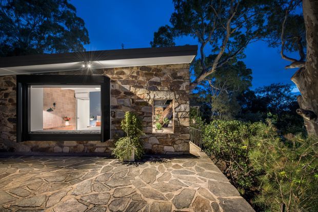 Twilight exterior view of a low, modern dwelling set among mature eucalyptus trees and native landscaping. The facade combines irregular natural stone walls with dark-framed glazing and a shallow low-pitched roof. A broad irregular stone-paved terrace leads toward the entrance, while the illuminated interior is visible through the large window.
