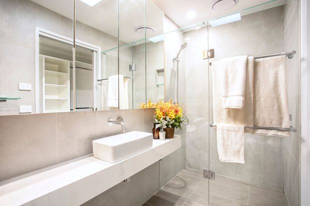 Bright contemporary bathroom with large-format light grey wall and floor tiles, a wall-mounted white vanity with integrated stone-look benchtop and vessel basin, frameless glass shower enclosure, mirrored wall, recessed shelving, and chrome towel rails. The space is clean, light, and compact, with a small floral arrangement adding colour.