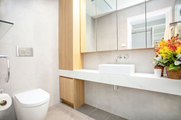 Bright contemporary bathroom featuring a wall-hung vanity with a white integrated benchtop and countertop basin, tall light-wood cabinetry, large mirrored storage cabinets, pale grey tiled walls, and grey floor tiles. A toilet and shower area are visible at the left, while a floral arrangement adds colour on the right.