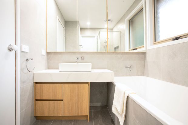 Bright, compact contemporary bathroom with a wall-mounted timber vanity, white rectangular basin, large mirrored splashback, freestanding-style bathtub, and pale stone-look wall tiles. A high horizontal window provides natural light, while chrome tapware and fittings add subtle contrast.