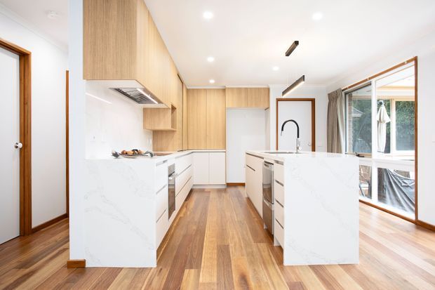 Bright contemporary kitchen with a galley-style layout and two white marble-look island benches. Light timber overhead cabinetry and matching timber flooring create warmth, while white lower cabinetry, integrated appliances, a black gooseneck tap, recessed ceiling lights and linear pendant lighting provide a clean modern finish. Large windows and a glazed opening bring in abundant natural light, with a timber-framed doorway visible at left.