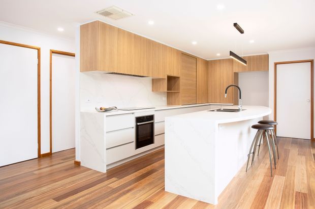 Bright contemporary kitchen with a large white stone island, integrated cooktop and sink, timber-grain overhead and wall cabinetry, white marble-look splashback and cabinetry panels, timber flooring, and minimalist black pendant lighting. The space includes a compact breakfast seating area with two stools and surrounding white doors with timber trim.
