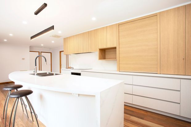 Bright contemporary kitchen with a large white waterfall island, integrated double sink and gooseneck tap, timber cabinetry, white drawers, timber flooring, and minimalist black accents. Two stools sit at the island, with a dining area and glazed timber-framed doors visible beyond.