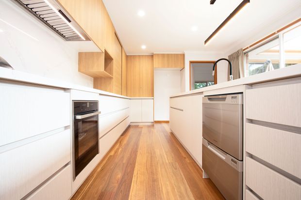 Bright contemporary galley kitchen with white handleless cabinetry, warm timber veneer wall cabinetry, integrated oven, stainless-steel dishwasher, pale benchtops and a central island with a black gooseneck tap. Warm timber flooring, linear recessed lighting and large windows create an airy, streamlined space.