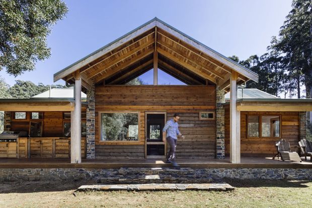 Front exterior view of a single-storey timber-clad house with a prominent gabled, covered entry porch. The facade combines horizontal natural timber boards, exposed timber soffits and posts, and dark rough-stone masonry piers and base walls. Large glazed openings and a central timber-framed entry door face a broad paved terrace, with side wings extending beneath lower pitched roofs. The setting is leafy and wooded, and the overall design has a rustic lodge or contemporary cabin character.