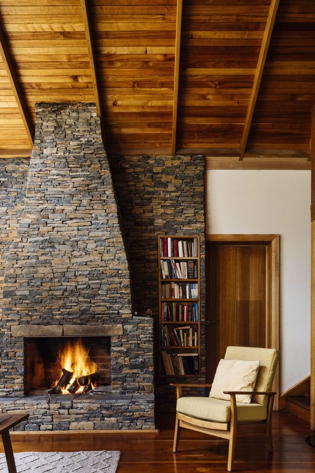 A warm, rustic interior centered on a substantial stacked-stone fireplace with a lit open fire. The room features a high exposed timber ceiling with visible rafters, dark timber flooring, timber doors and trim, a built-in bookcase, and a mid-century-style upholstered armchair. The palette is rich in brown wood, grey-brown stone, cream upholstery, and warm firelight.