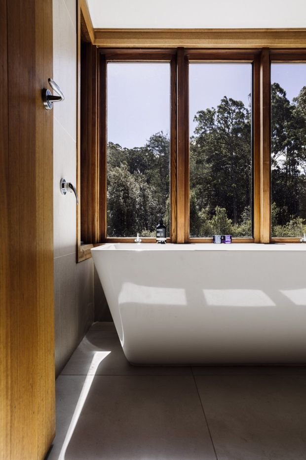 A contemporary bathroom featuring a freestanding white bathtub positioned beneath a bank of large timber-framed windows. The room has dark large-format floor tiles, pale wall surfaces, warm wood detailing, and abundant natural light with views of trees outside. A timber door with chrome hardware is visible at left.