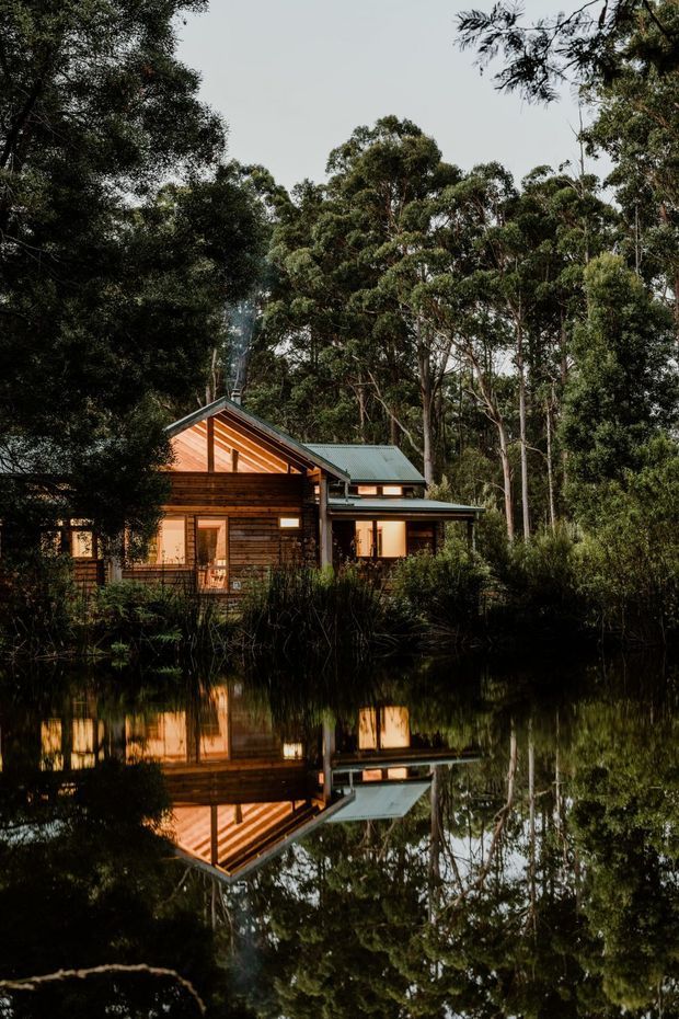 A timber-clad house or cabin set within dense eucalyptus woodland, viewed across a still pond that creates a strong reflection of the building and surrounding trees. The dwelling has a pitched gabled roof, dark metal roofing, warm horizontal timber boards, large windows, and a connected lower-pitched wing. The scene is an atmospheric exterior photograph with natural vegetation and subdued evening-like lighting.