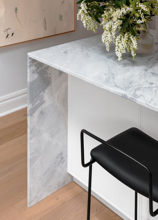Close-up interior detail of a minimalist stone-topped built-in counter or peninsula with a waterfall end panel, paired with a black bar stool. The counter features prominent pale grey and white veined stone, with white cabinetry beneath, light timber flooring, and a soft neutral wall. A vase of white flowers and framed artwork add restrained decorative elements.