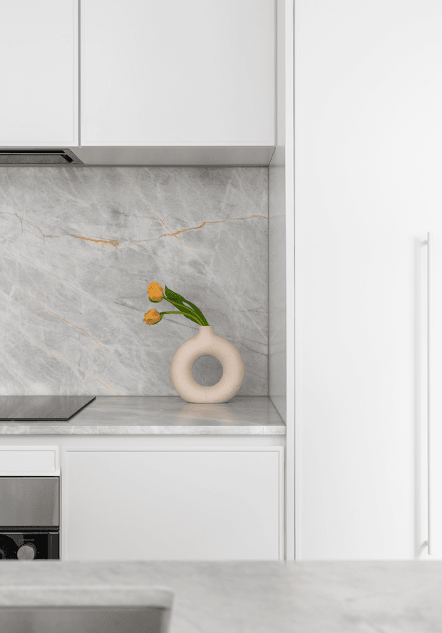Close-up view of a refined contemporary kitchen, showing white flat-panel cabinetry, a grey marble-look splashback and benchtop with subtle gold veining, integrated stainless-steel appliances, and a small sculptural ceramic vase on the counter.