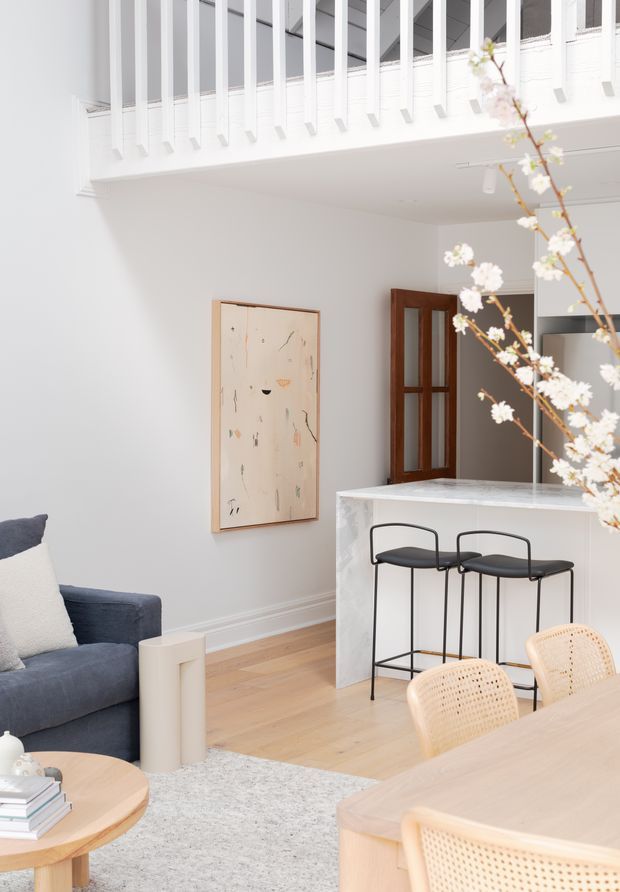 Bright, refined open-plan living and dining area with a compact marble-look kitchen island and two black bar stools. The room features white walls, light timber flooring, a white balustraded upper-level void or mezzanine, a dark blue sofa, pale timber dining furniture with woven cane chairs, and a warm timber-framed glazed door. A flowering branch is visible in the foreground.