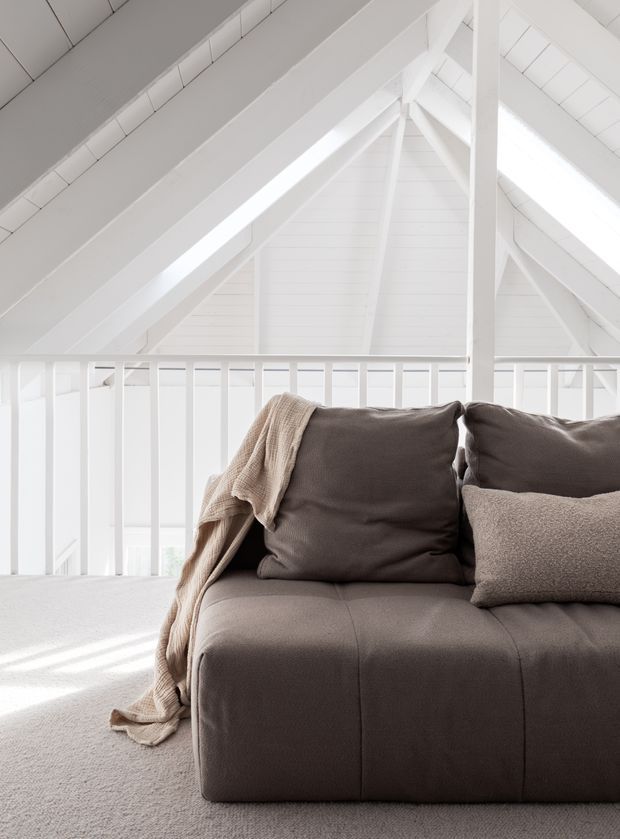 Bright, minimalist loft-like interior beneath a steep white pitched roof, with exposed painted timber rafters and a white balustrade overlooking a lower level. A large taupe-grey upholstered sofa with neutral cushions and a draped beige throw occupies the foreground, set on a pale carpet. Strong natural light creates an airy, calm atmosphere.