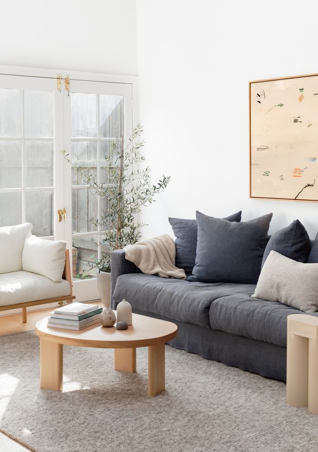Bright, softly styled living room with a deep charcoal-blue sofa, cream armchair, round light-wood coffee table, textured neutral rug, and a small olive tree beside white French doors. A framed abstract artwork hangs above the sofa. The room has abundant natural light and a calm, neutral palette with understated Scandinavian-influenced furnishings.