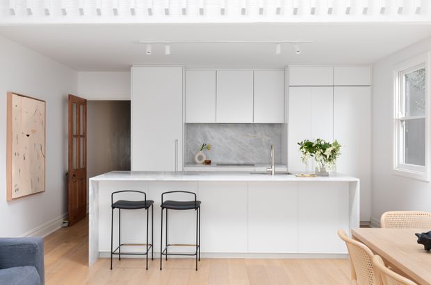 Bright, compact open-plan kitchen with a large white island, integrated white cabinetry, pale marble-look splashback, timber flooring, and two black bar stools. A dining area with woven chairs is visible at right, while a living area is partially visible at left. The room has white walls, abundant daylight, a skylight or rooflight detail, and a clean, refined contemporary aesthetic.