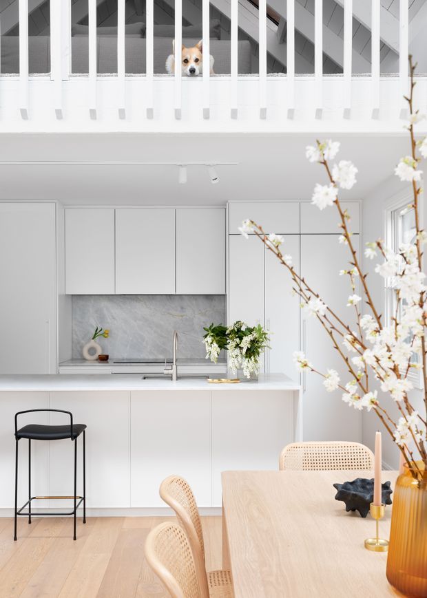 Bright, predominantly white open-plan kitchen and dining area with a mezzanine or loft level above. The kitchen has flat-panel white cabinetry, a pale veined stone backsplash and benchtop, an island with an undermount sink and brushed metal tap, and light timber flooring. A pale timber dining table with woven cane chairs occupies the foreground, while a black minimalist bar stool sits at the island. White balustrade rails and a small dog are visible on the upper level. Natural light enters through large windows, with floral branches and simple decorative objects adding softness.