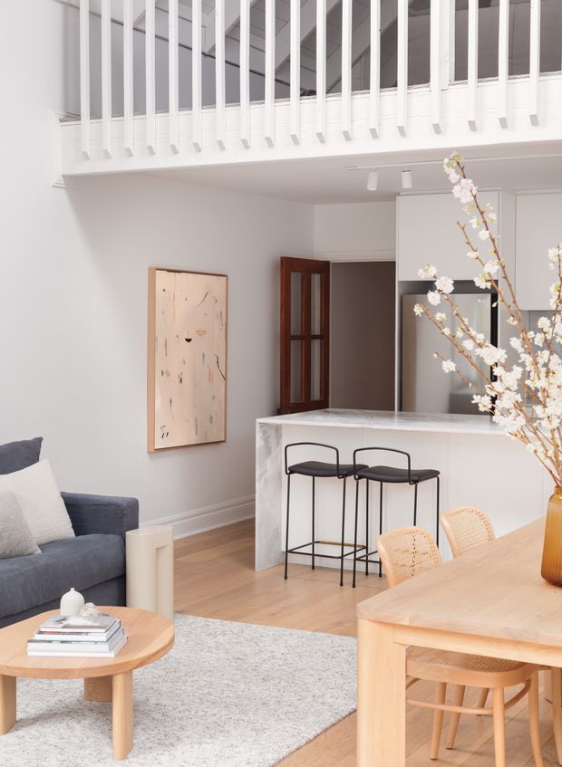 Bright, airy open-plan interior combining a living room, dining area and kitchen. The room features white walls and cabinetry, a marble-look kitchen island with dark bar stools, pale timber flooring, a light timber dining table with woven chairs, a blue-grey sofa, and a soft neutral rug. A white balustrade overlooks the space, suggesting a mezzanine or upper-level landing. Natural light and restrained furnishings create a calm contemporary atmosphere.