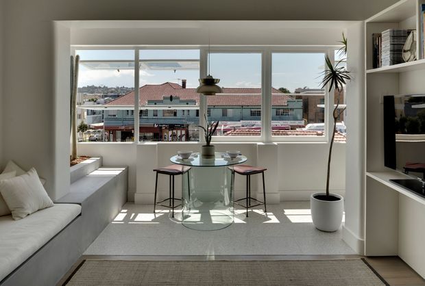 Bright, compact open-plan living and dining area with a built-in white window seat, white sofa, round glass dining table, and two stools. Floor-to-ceiling-style windows provide abundant daylight and views toward neighbouring buildings. White built-in shelving and media joinery line the right side, while potted plants add greenery. The space has a clean, restrained contemporary appearance with pale finishes and strong natural light.