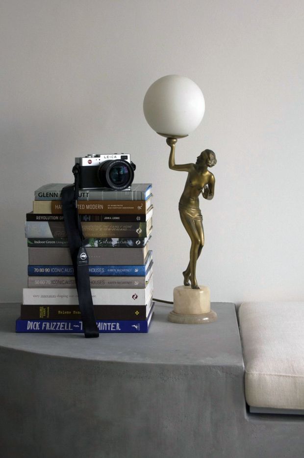 A minimalist interior vignette featuring a stack of design and photography books with a Leica camera, beside a sculptural brass-toned figurine lamp with a white globe shade. The objects sit on a smooth grey concrete or plaster-finished surface next to a light upholstered sofa, against a plain warm-grey wall.