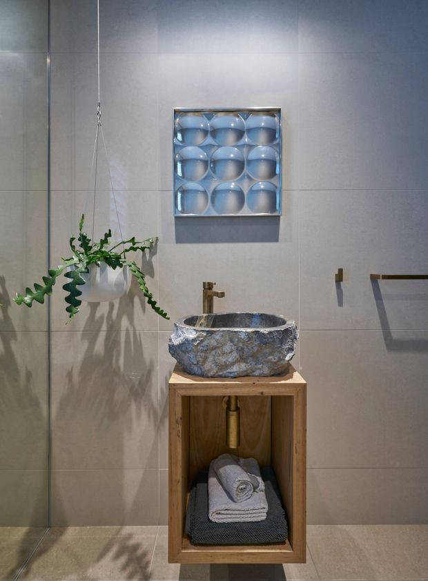 A compact, refined bathroom featuring large grey-beige tiled walls and floor, a rough natural stone vessel basin on an open timber vanity, and a brushed brass-toned wall-mounted tap. A hanging plant adds greenery, while a square decorative wall artwork and simple towel rail complete the space. The room has a warm, earthy, softly lit character with a strong natural-material focus.