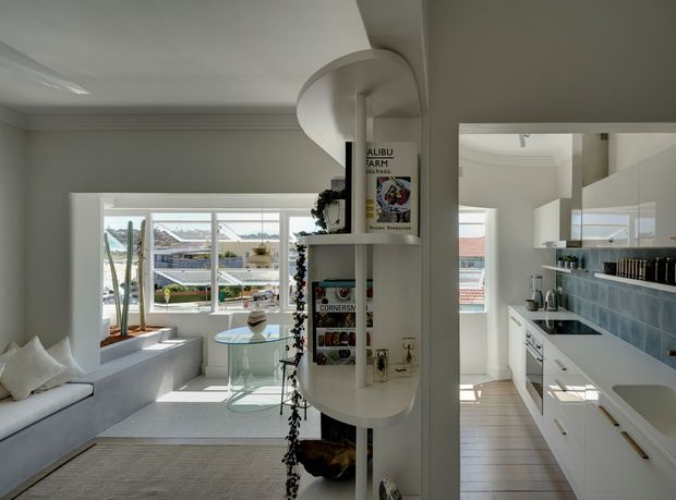 Bright open-plan residential interior showing a compact white kitchen on the right and a sunlit sitting or living area on the left. A curved built-in shelving divider separates the spaces, with integrated display storage and trailing greenery. The room has pale walls and cabinetry, large windows, a tiled kitchen splashback, light timber flooring, and a built-in window seat overlooking neighbouring rooftops.