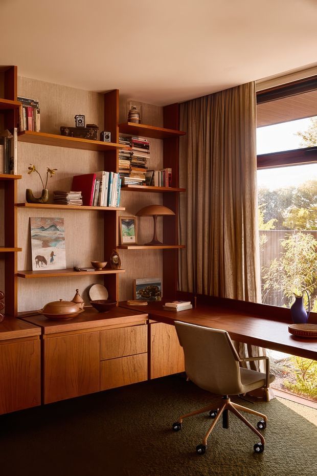 Warm, mid-century-inspired home office with extensive built-in timber shelving and cabinetry, a long integrated desk beneath a large timber-framed window, curated books and ceramics, and a vintage-style upholstered swivel chair. The room has textured neutral wallcovering, full-height brown curtains, timber joinery, and a dark carpet, with abundant natural light and garden views through the window.