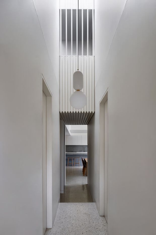 A narrow, minimalist residential hallway with pale walls and terrazzo flooring, framed by doorways on both sides. A dramatic double-height void is crossed by a slatted overhead bridge or screen, with two suspended spherical pendant lights. The corridor leads toward a contemporary kitchen and dining area in the distance.
