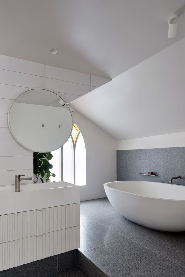 A bright, refined contemporary bathroom with a freestanding white bathtub, wall-mounted white vanity, large round mirror, and brushed metal tapware. Pale linear wall tiles and a textured grey feature wall contrast with dark grey floor tiles. Arched windows bring in natural light, while a small plant adds organic detail.