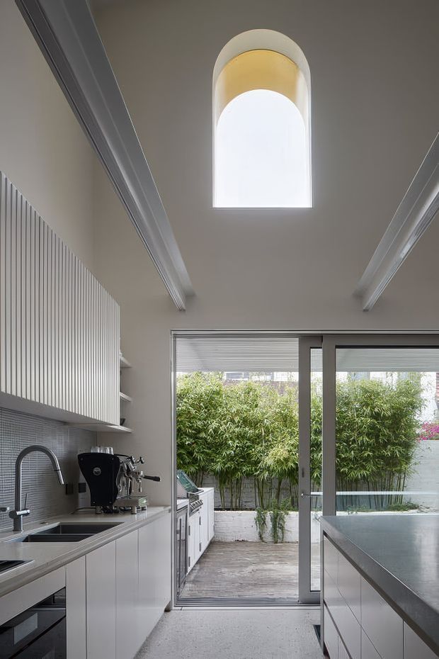 A narrow, contemporary kitchen viewed toward a glazed opening leading to a small planted courtyard. The kitchen has pale grey fixed cabinetry, a dark stone or composite benchtop, a stainless-steel sink and tap, a coffee machine, and a fine grey tiled backsplash. A large central island occupies the right foreground. The room features a high ceiling with exposed painted beams and a tall arched skylight or clerestory window, plus full-height sliding glazing and roller blinds.