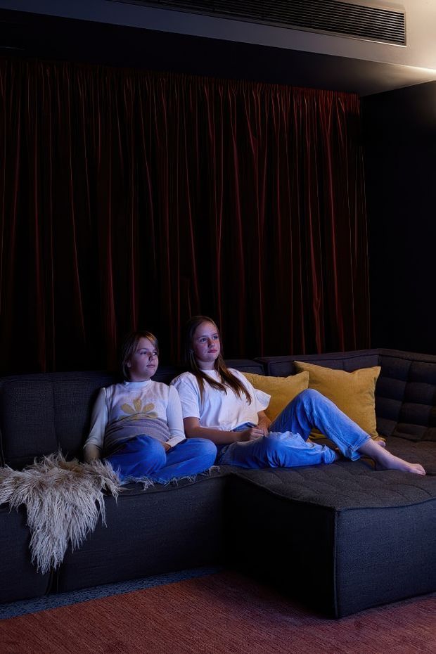 A dark, cozy home cinema or media room with two children relaxing on a large charcoal sectional sofa. The room features deep burgundy acoustic curtains, dark walls, warm mustard cushions, a textured throw, and a reddish-brown carpet. A linear ceiling air-conditioning vent is visible, and the low lighting creates an immersive screening-room atmosphere.