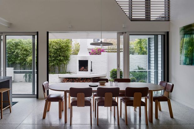 Bright contemporary dining room with a long oval timber dining table and mid-century inspired upholstered chairs. Full-height black-framed glazing opens views to a planted courtyard, while a white masonry fireplace and suspended circular pendant light form the central backdrop. A louvred skylight or clerestory feature adds overhead interest, and polished concrete flooring reinforces the clean modern character.