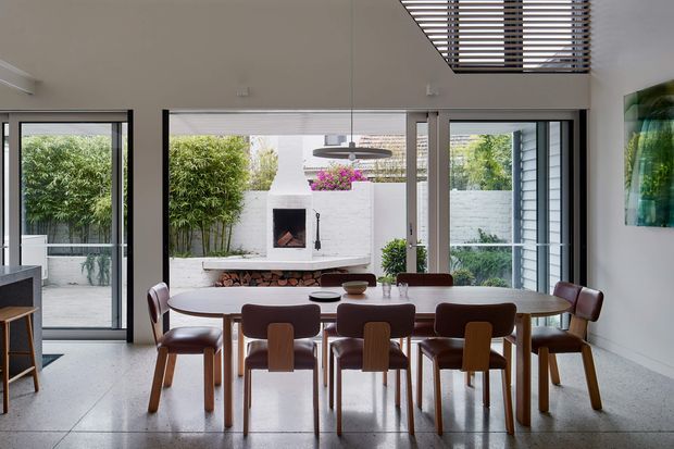 A bright contemporary dining area with a large oval timber dining table and numerous upholstered timber dining chairs. The room has polished grey concrete flooring, white walls, extensive black-framed glazing and sliding doors, and a partially visible kitchen at left. A white brick fireplace with a firebox forms the focal point beyond the table, opening to a leafy courtyard. Horizontal timber louvres or blinds are visible high on the ceiling, while a sculptural pendant light hangs above the dining table.