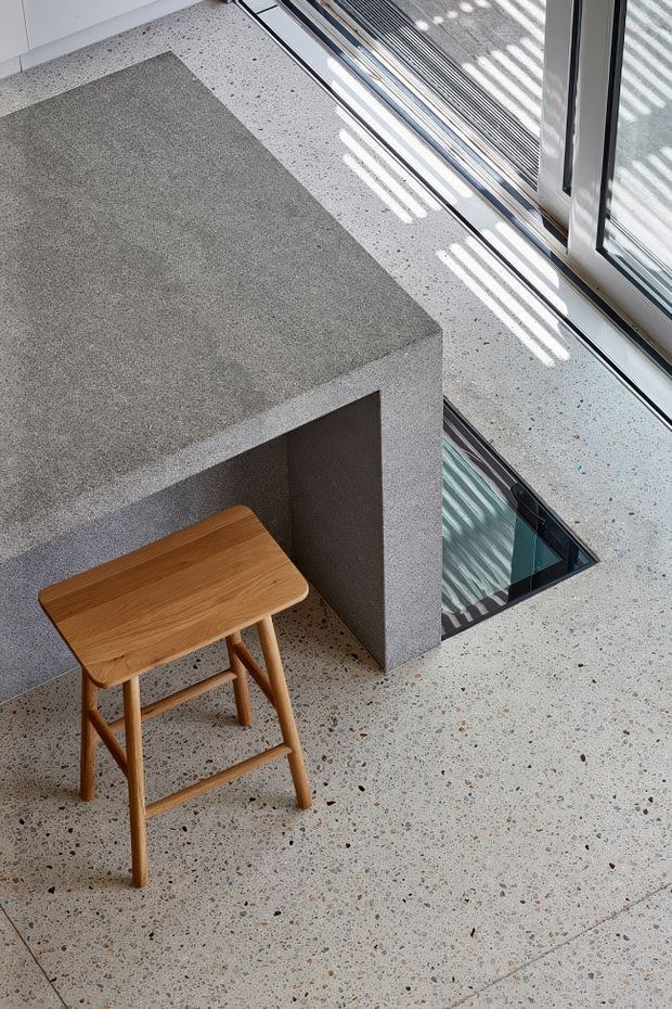 A bright, contemporary interior detail showing a large grey textured built-in counter or island with an open underside, a simple light timber stool, and a speckled terrazzo floor. Full-height glazed doors or windows line the right side, casting striped sunlight across the floor and surfaces. A small glazed floor opening or skylight is visible beside the counter.