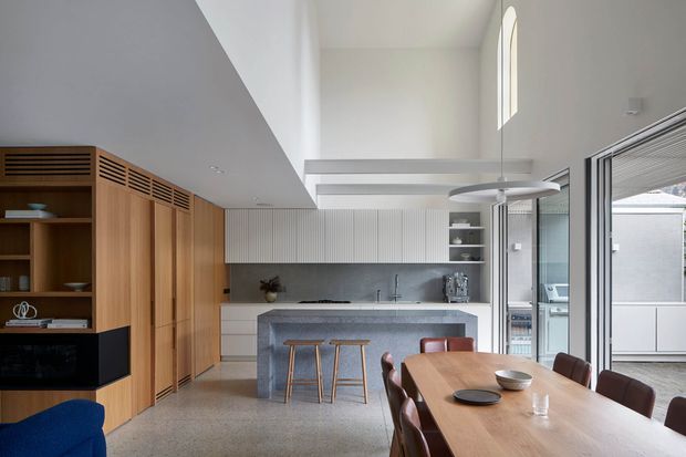 Bright contemporary open-plan kitchen and dining area with a high ceiling and clerestory-style arched window. The kitchen features extensive vertical timber cabinetry, white ribbed upper cabinetry, a dark grey stone-look benchtop and splashback, and an island with timber stools. A large timber dining table with brown upholstered chairs occupies the foreground. Glazed sliding doors open to an outdoor area, while a sculptural circular pendant hangs above the dining zone.