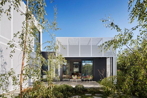 A contemporary multi-level building viewed from a landscaped garden courtyard. The dark-framed glazed entry and covered outdoor seating area are enclosed by distinctive white perforated metal screens, with planted greenery in the foreground and clear blue sky above.