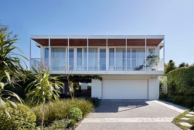 A modern two-storey residential facade viewed from the driveway, featuring a broad upper-level balcony with slim white metal balustrades, extensive glazed doors, a timber-lined soffit, and a white rendered lower level with an integrated double garage. The landscaped front garden includes dense screening hedges, ornamental grasses, tropical foliage, and a paved driveway and path.