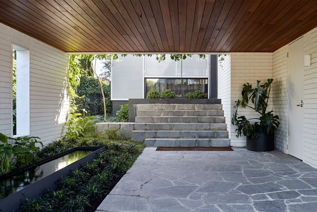 Covered residential entry courtyard with white horizontal wall cladding, a warm timber-lined ceiling, irregular grey stone paving, and broad grey stone steps leading to a glazed entrance. Lush planting and a narrow reflecting pond frame the approach, while a modern neighbouring or internal facade with perforated screening is visible beyond.