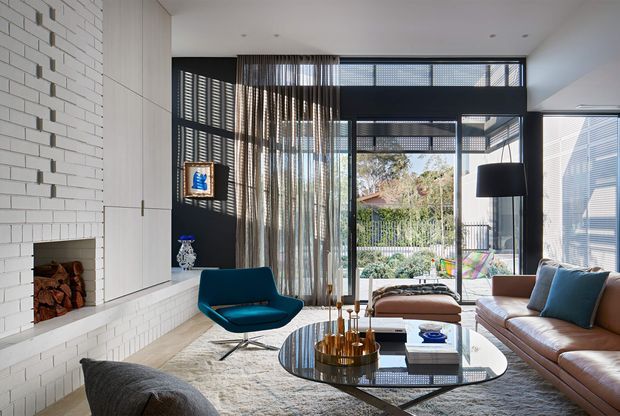 Bright contemporary living room with a white brick feature fireplace, dark-framed floor-to-ceiling glazing, sheer curtains, a brown leather sofa, blue accent chair, glass coffee table, and views to an outdoor terrace and garden. The room combines strong natural light with dark architectural framing and textured furnishings.