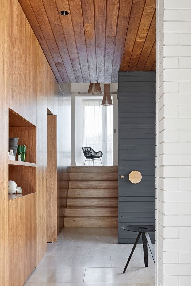 A vertically framed contemporary interior hallway and stair landing. Warm timber wall panelling, open built-in shelving, and a richly stained timber plank ceiling contrast with a dark charcoal slatted door or partition and a white painted brick wall. Broad timber steps lead to a bright upper landing with a black sculptural chair and pendant lights. Pale stone or concrete-look floor tiles and restrained furnishings create a refined, tactile composition.