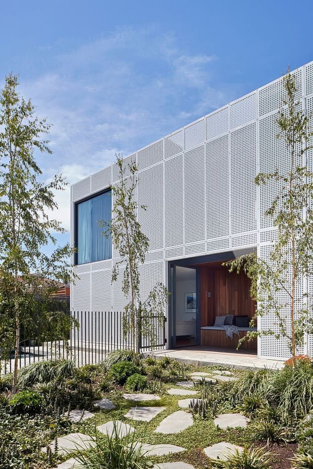 Exterior view of a contemporary two-storey residence with a rectilinear form and extensive white perforated screening across the facade. A large glazed opening and recessed entry or living area reveal warm timber lining and built-in seating. The building is set behind a landscaped front garden with irregular concrete stepping stones, grasses and low planting.