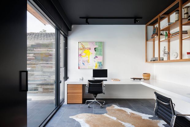 A contemporary home office with a long white L-shaped built-in desk, two black office chairs, computer workstation, timber drawer unit, open timber shelving, and a colourful abstract artwork. The room has a dark ceiling with track lighting, large black-framed glazing, dark grey tiled flooring, and a cowhide rug. An exterior tiled wall is visible through the glazing.