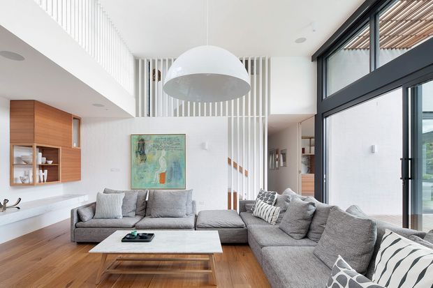 Bright contemporary open-plan living room with a large grey sectional sofa, timber flooring, a low light-coloured coffee table, and built-in timber wall cabinetry. A double-height void with a sculptural pendant, vertical white architectural screening, stair access, and extensive dark-framed glazing create an airy, refined interior.